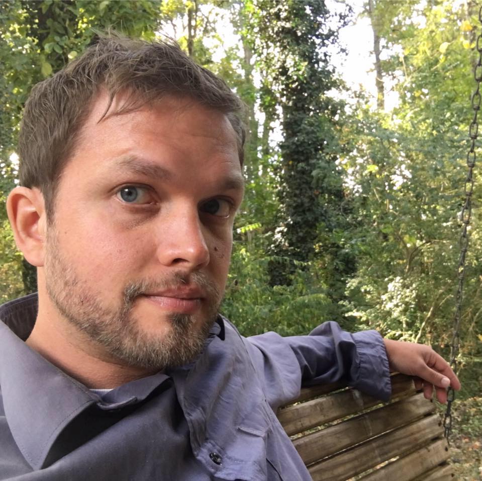 Eric Moore, a 30-something man with light beard looks 3/4 toward the camera, sitting on bench in wooded area.